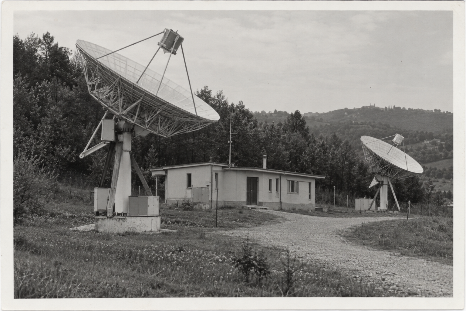 Fotografia in bianco e nero di due parabole radioastronomiche, un basso edificio tecnico e la strada di accesso fra le colline.